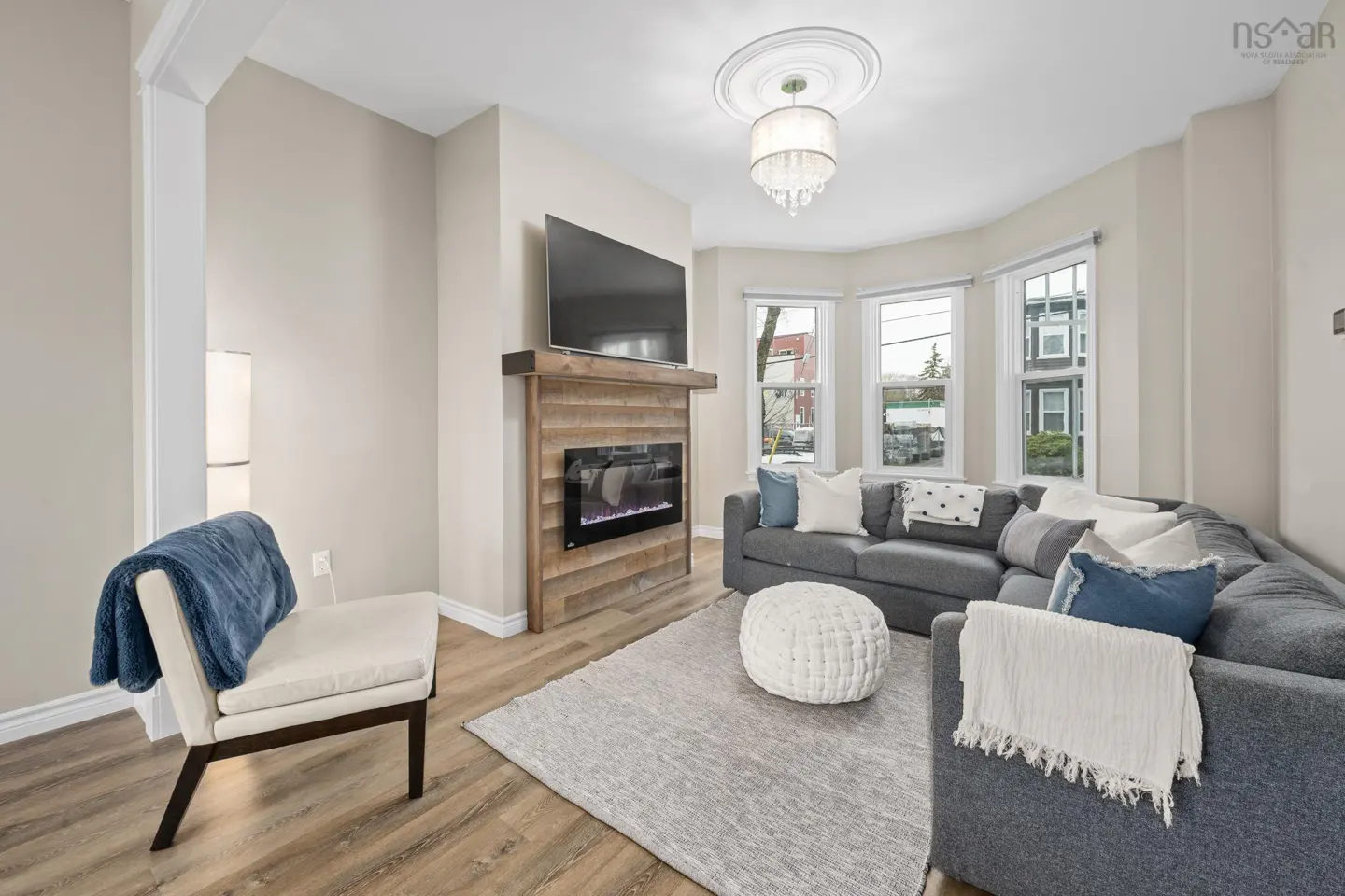 Living room with gray sectional, white ottoman, and a fireplace with a TV above it. A chair with a blue blanket is on the left.