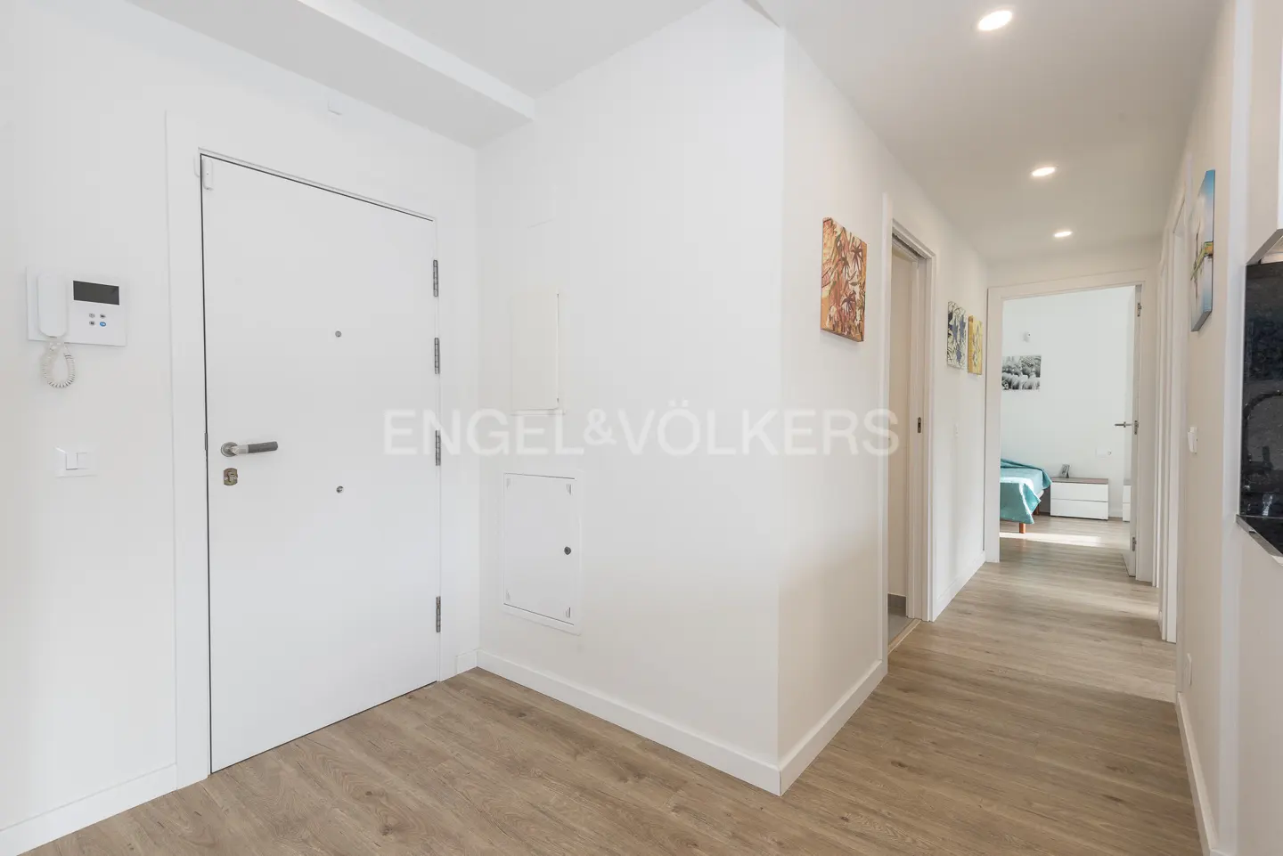 Apartment entryway with a white door, intercom, and light wood floors. A hallway leads to a bedroom with a blue bedspread.