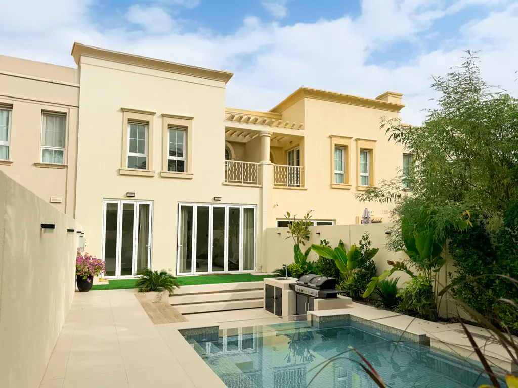 Backyard view of a tan two-story house with a pool, grill, and patio. Blue sky with clouds above.