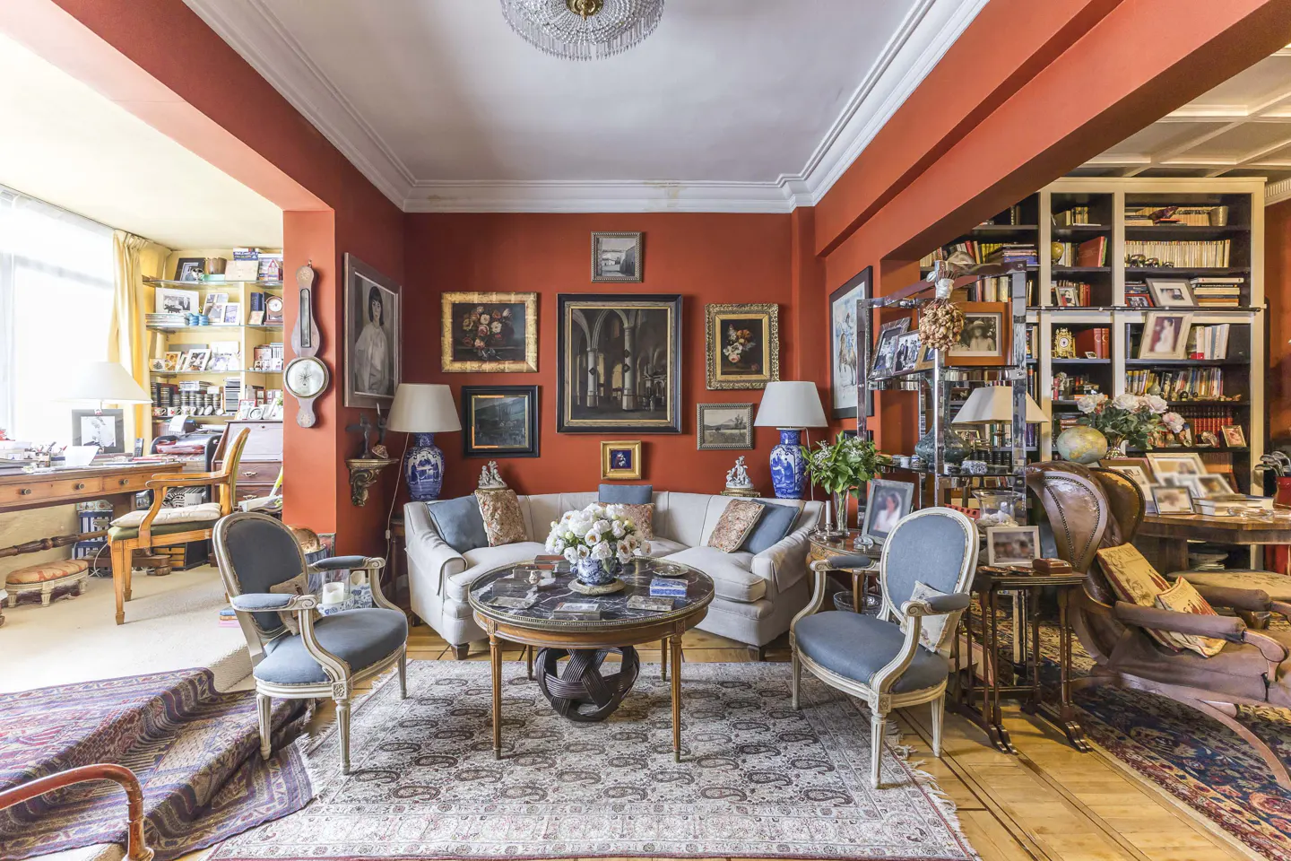 Eclectic living room with orange walls, white sofa, blue chairs, and a round table on a patterned rug. Artwork adorns the walls.