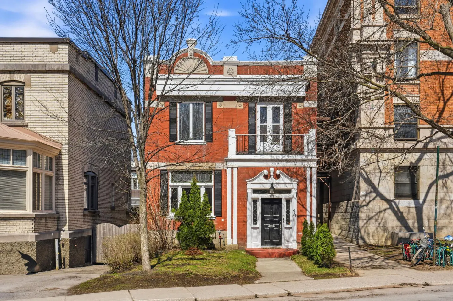 Two-story red brick house with white trim, black shutters, and a small balcony. Bare trees frame the building against a blue sky.