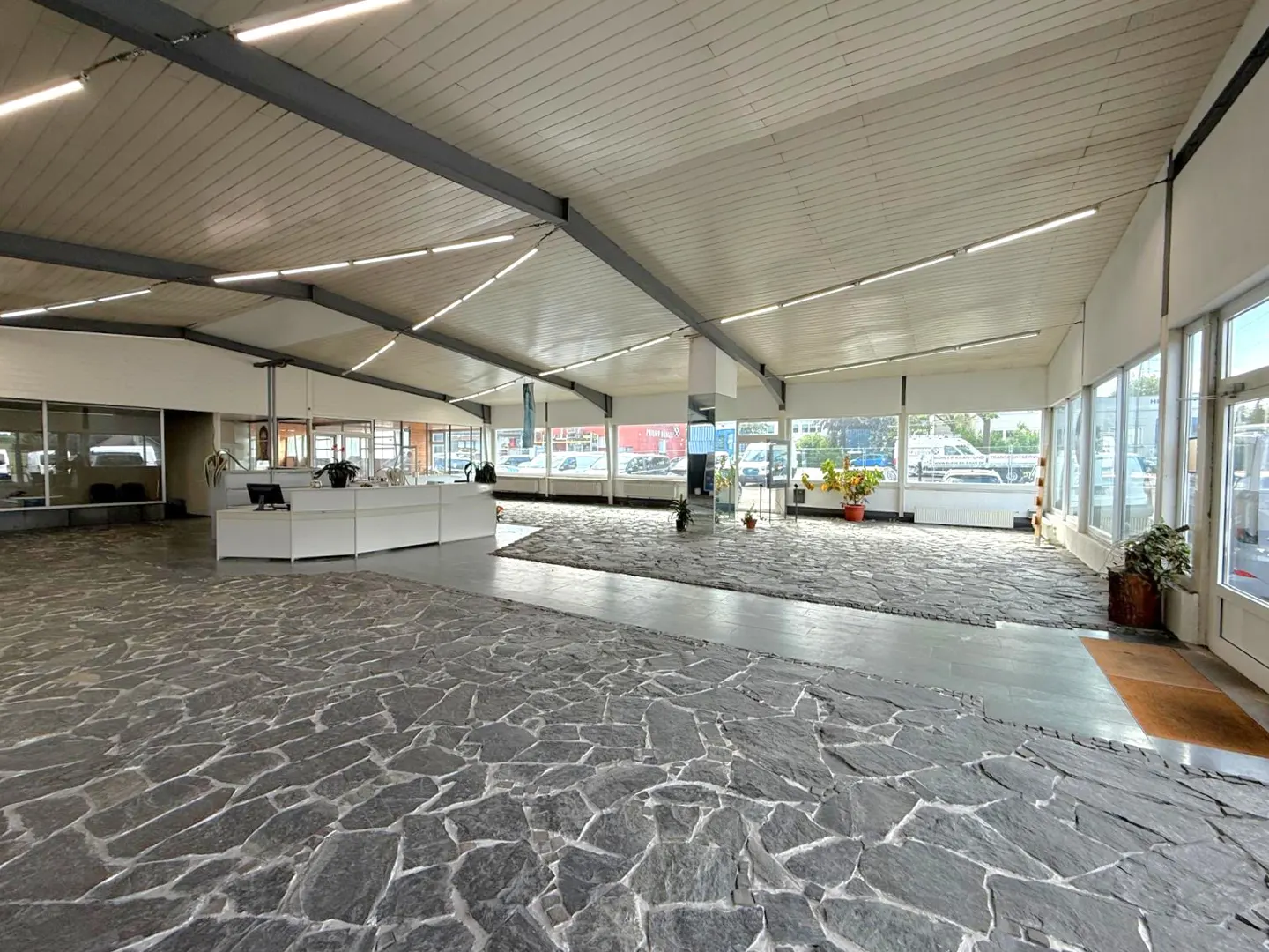 Wide shot of a car dealership interior with stone floors, white walls, and large windows. A white reception desk is visible.