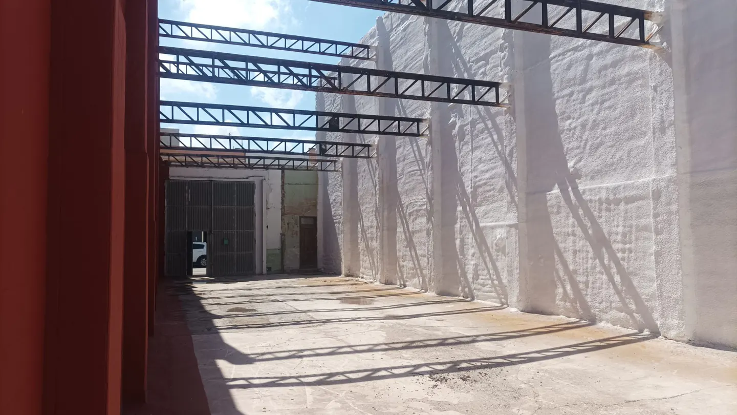 A warehouse interior with metal beams casting shadows on the concrete floor and white walls. A red column is on the left.