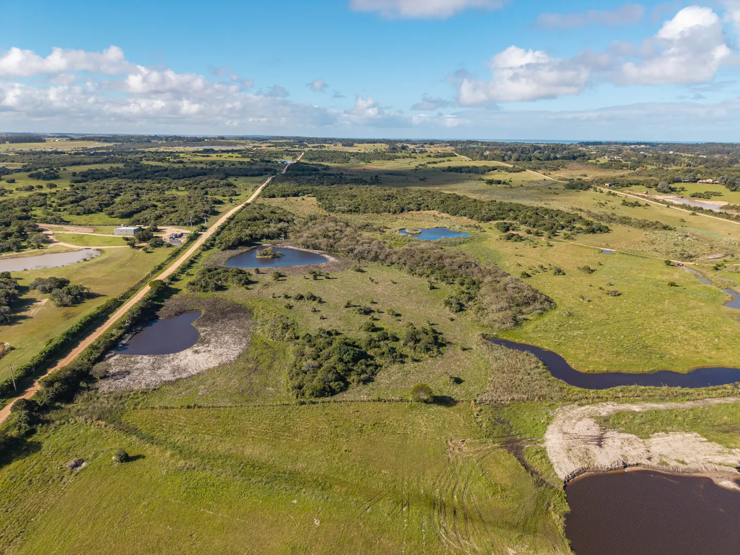 Aerial view of green fields with ponds, trees, and a dirt road under a blue sky with scattered clouds.