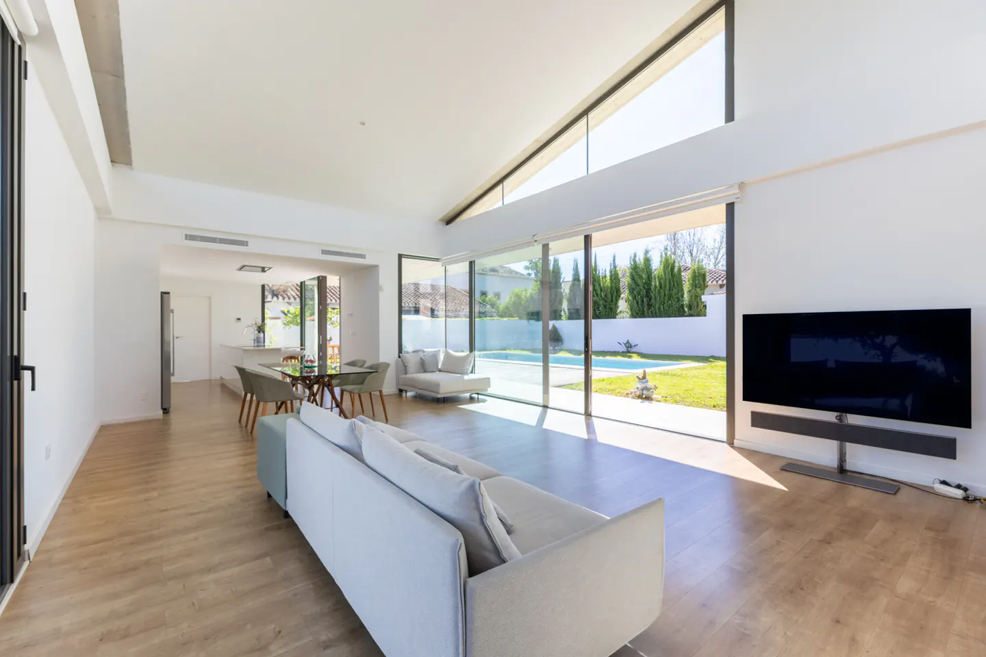 Bright, modern living room with white walls, light wood floors, and large windows overlooking a pool and backyard. A white sofa faces a TV.