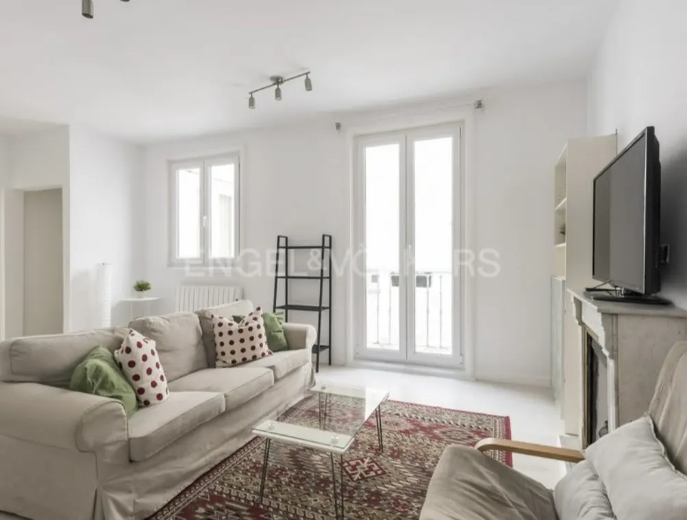 Bright living room with white walls, a beige sofa with patterned pillows, and a patterned rug. A TV sits on a mantelpiece.