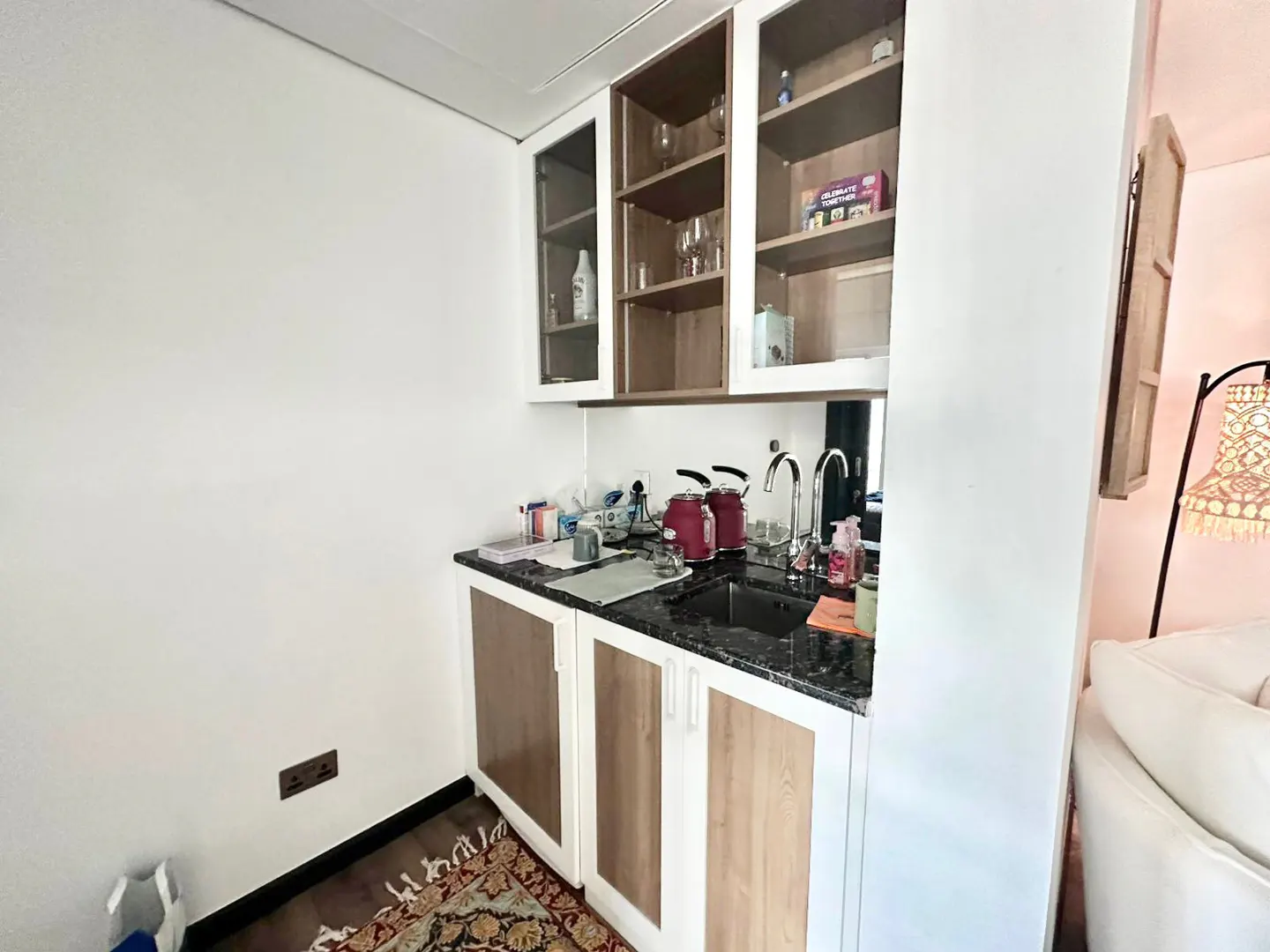 A small kitchen area with white cabinets, a black countertop, and a sink. There are shelves above the counter with glasses and other items. A red kettle sits on the counter.