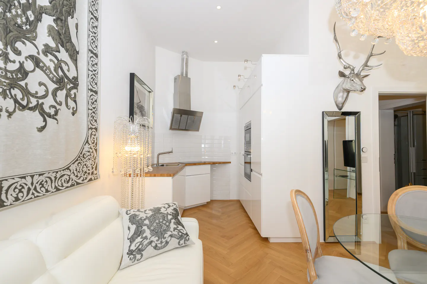 A bright, modern apartment interior with white walls, a white sofa, and a small kitchen area. A decorative tapestry hangs on the wall.