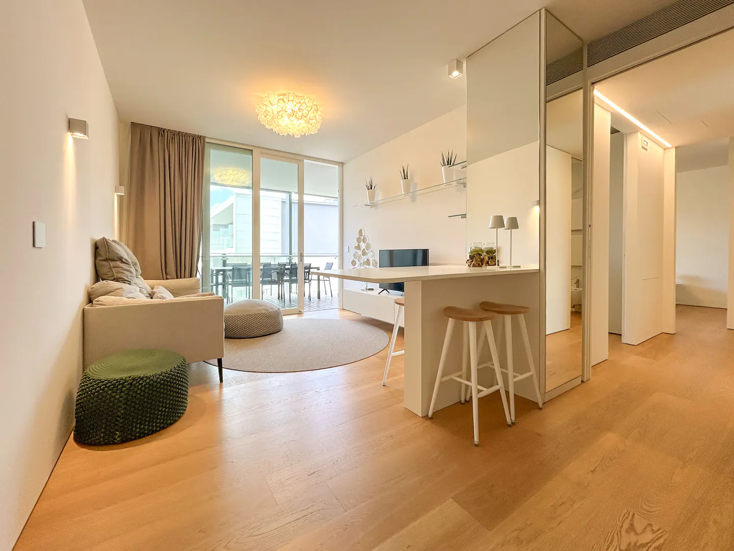 Bright, modern apartment interior with wood floors, a beige sofa, and a white kitchen island with two wooden stools. A large glass door leads to a balcony.