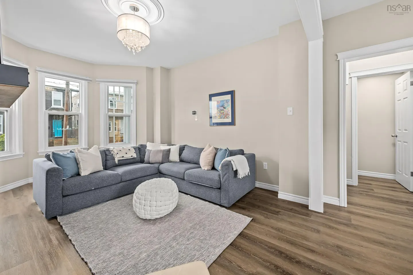 Living room with a gray sectional sofa, white ottoman, and gray rug on a wood floor. Windows look out onto a street. A chandelier hangs from the ceiling.