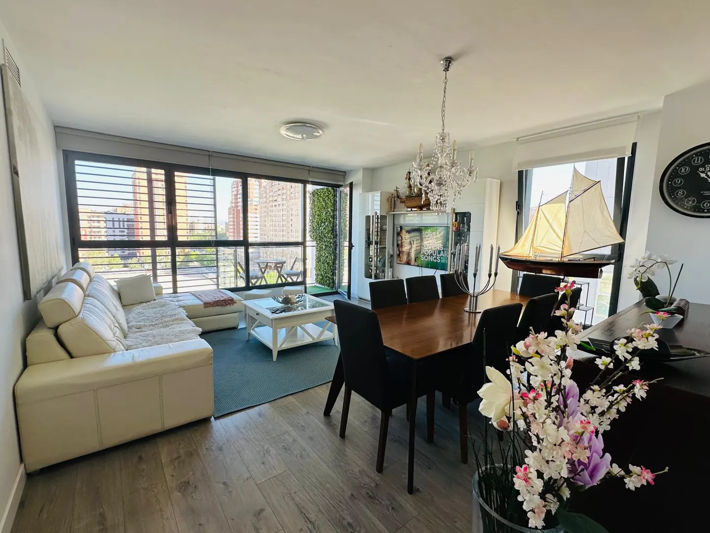 Bright living room with a white couch, wood floors, and a dining table with black chairs. A model sailboat sits near a window.