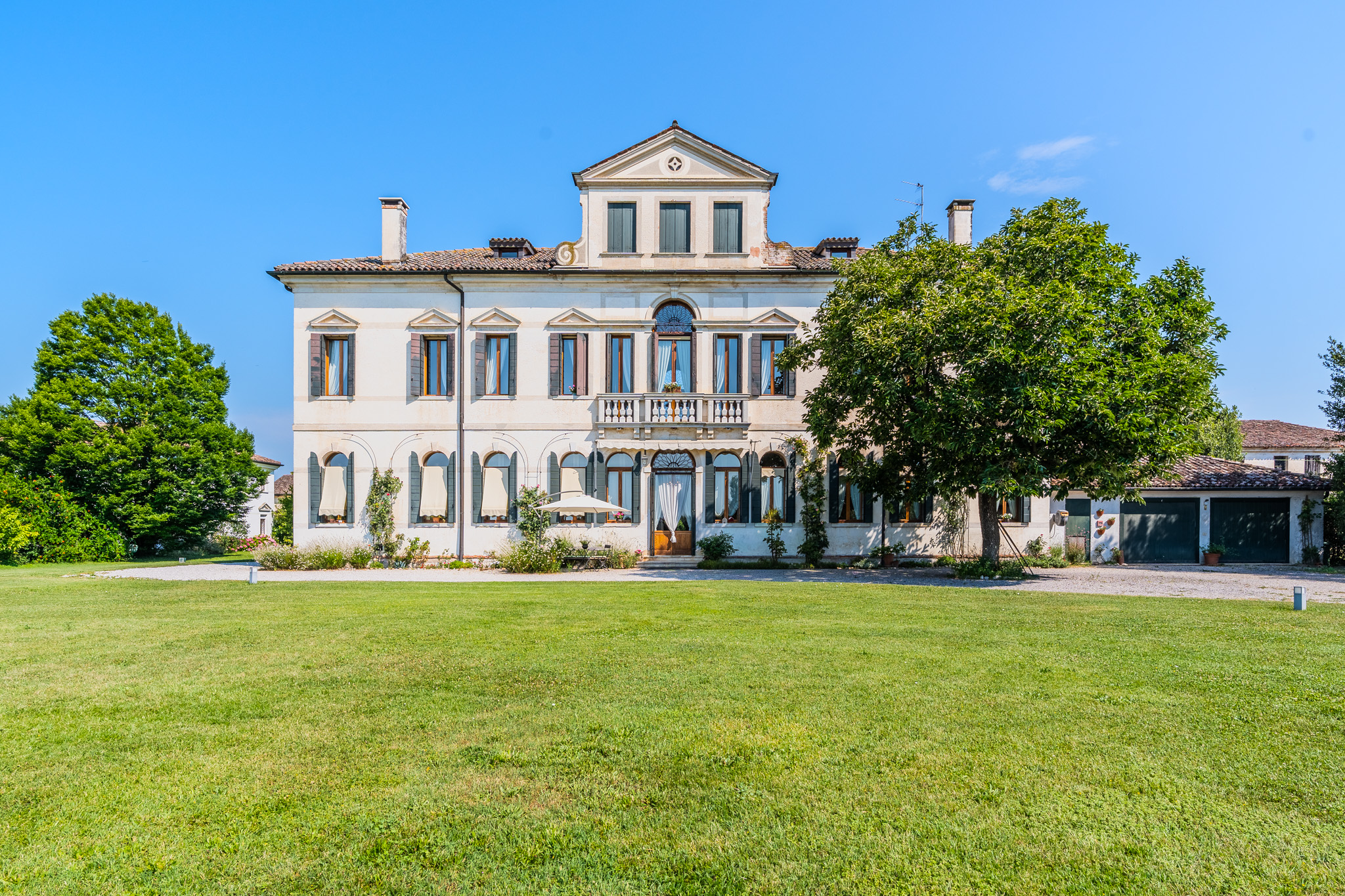 A grand, three-story cream-colored villa with dark shutters, a balcony, and a large green lawn under a clear blue sky.