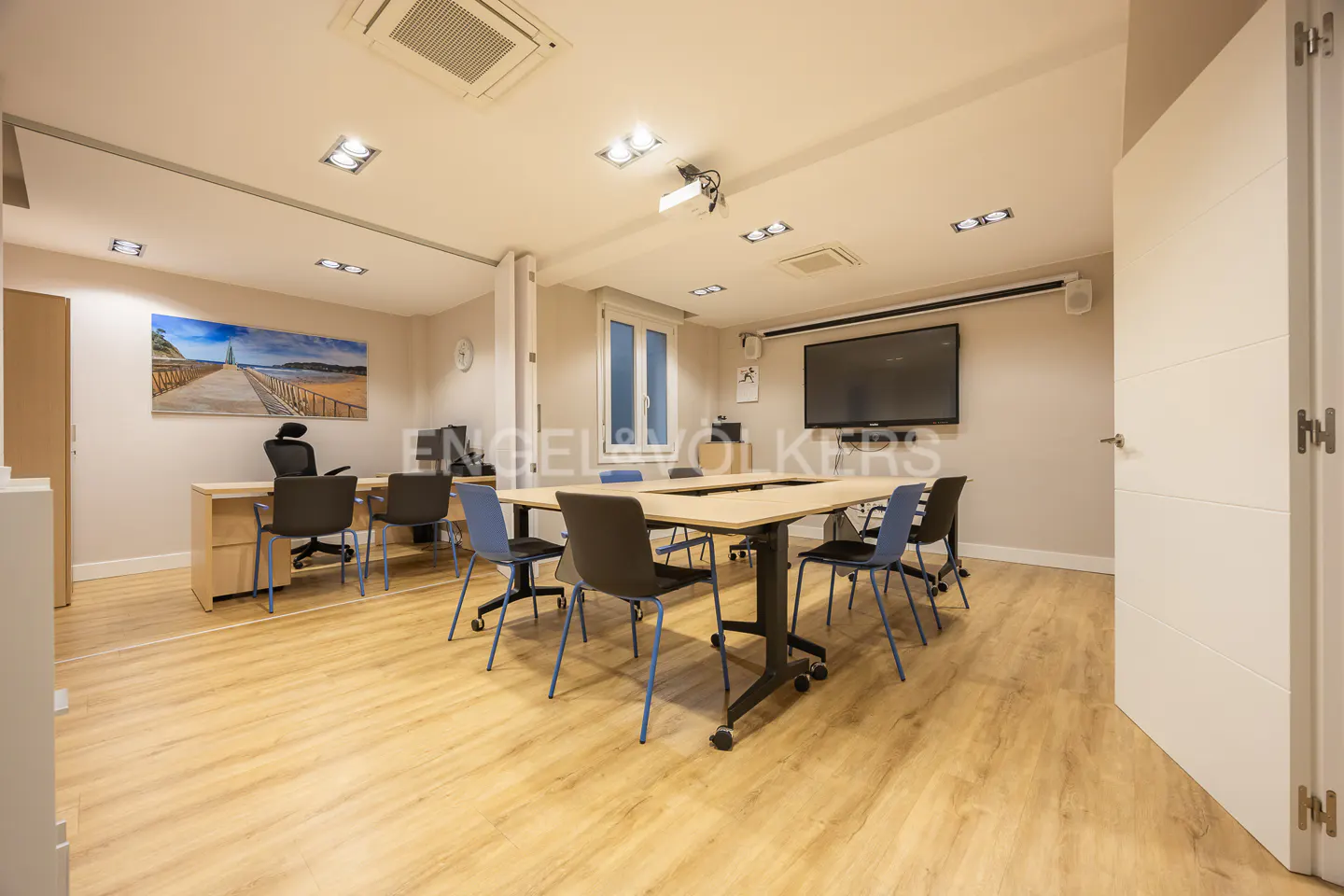 Bright office space with a U-shaped table, black chairs, and a large screen TV on the wall. A beach scene hangs above a desk.