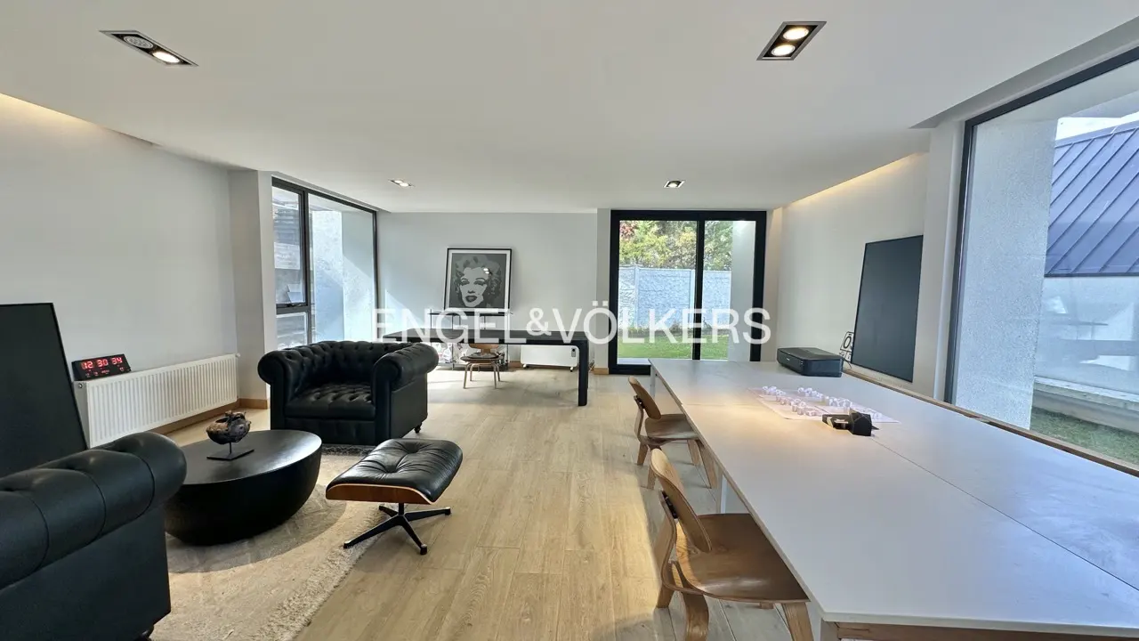 Bright, modern living room with black leather sofas, a long white table, and large windows overlooking a green yard.