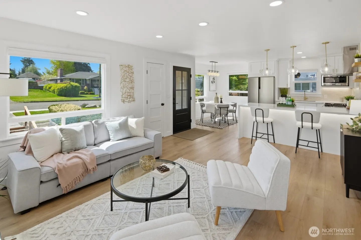 Bright, open-concept living space with a gray sofa, white chairs, and a modern kitchen with a breakfast bar. Large windows offer a view of a green lawn.