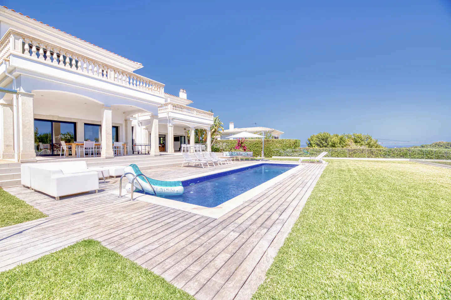 Luxury white villa with a swimming pool, wooden deck, white lounge furniture, and a large green lawn under a clear blue sky.
