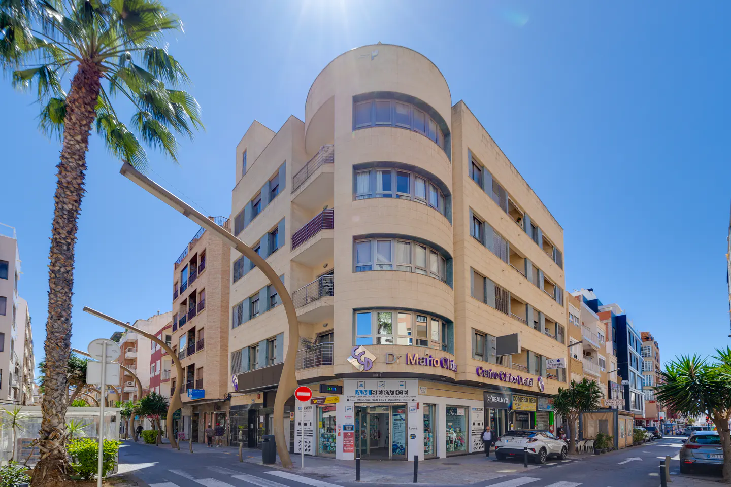 Street view of a tan building with shops on the ground floor, palm trees, and a blue sky.
