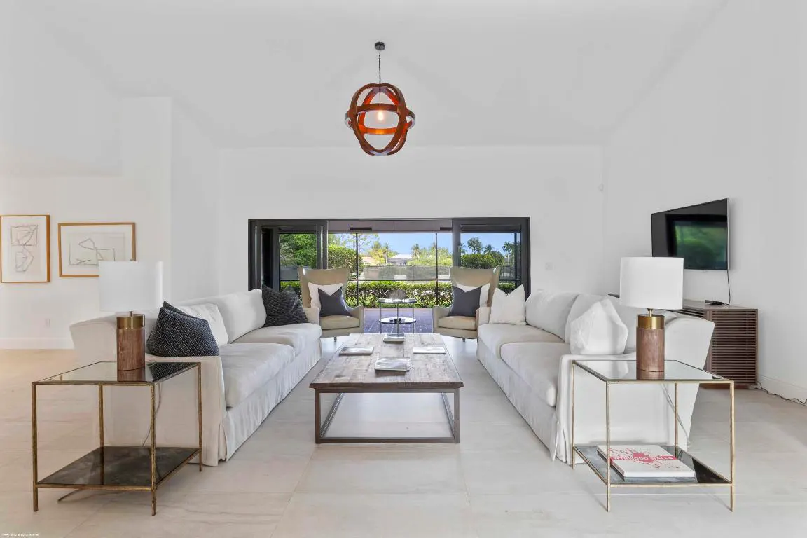 Bright living room with white sofas, wood coffee table, and glass side tables. A modern light fixture hangs from the ceiling. Patio view in the background.