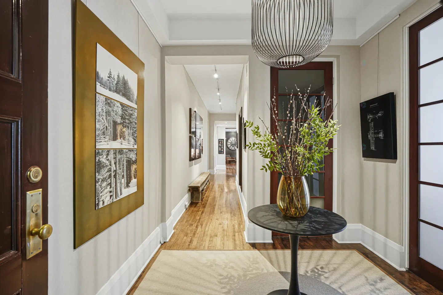 Hallway with hardwood floors, beige walls, and artwork. A round table with a vase of greenery sits near a window. A long bench lines the hallway.