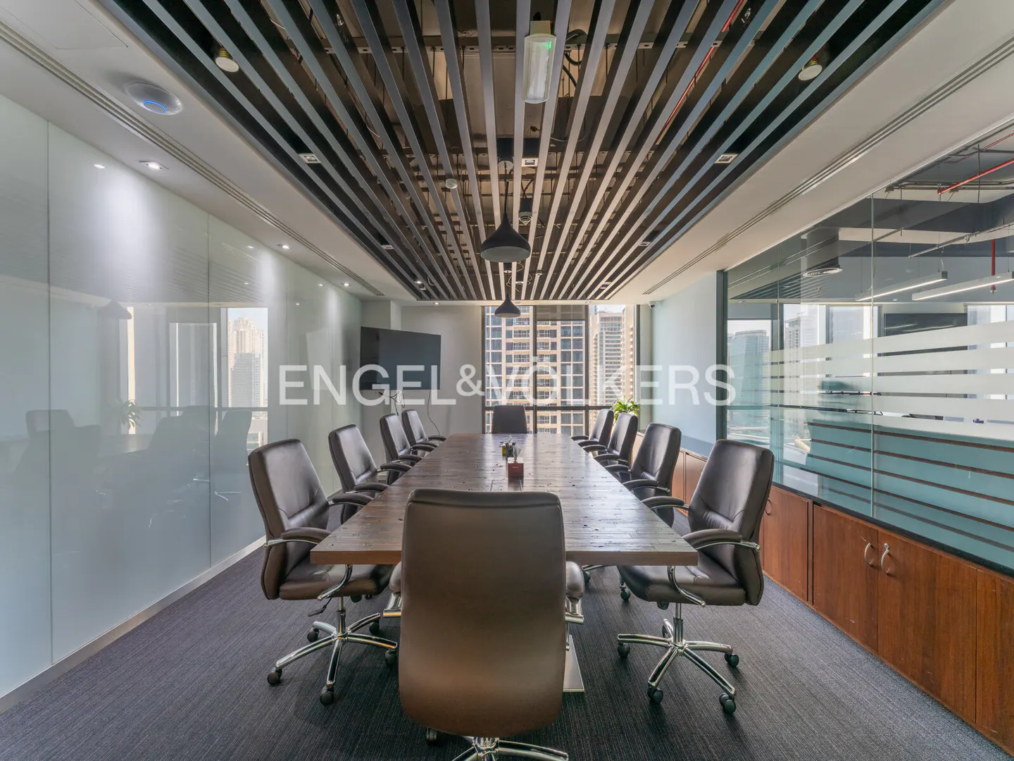 A modern conference room with a long wooden table, leather chairs, and a ceiling with metal slats. The Engel & Volkers logo is on a glass wall.