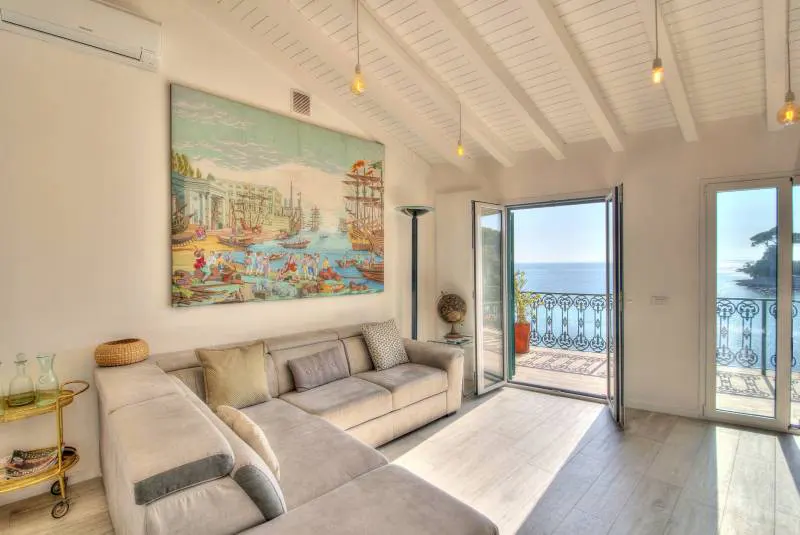 Living room with a gray sectional sofa, a large painting, and an open balcony overlooking the ocean.