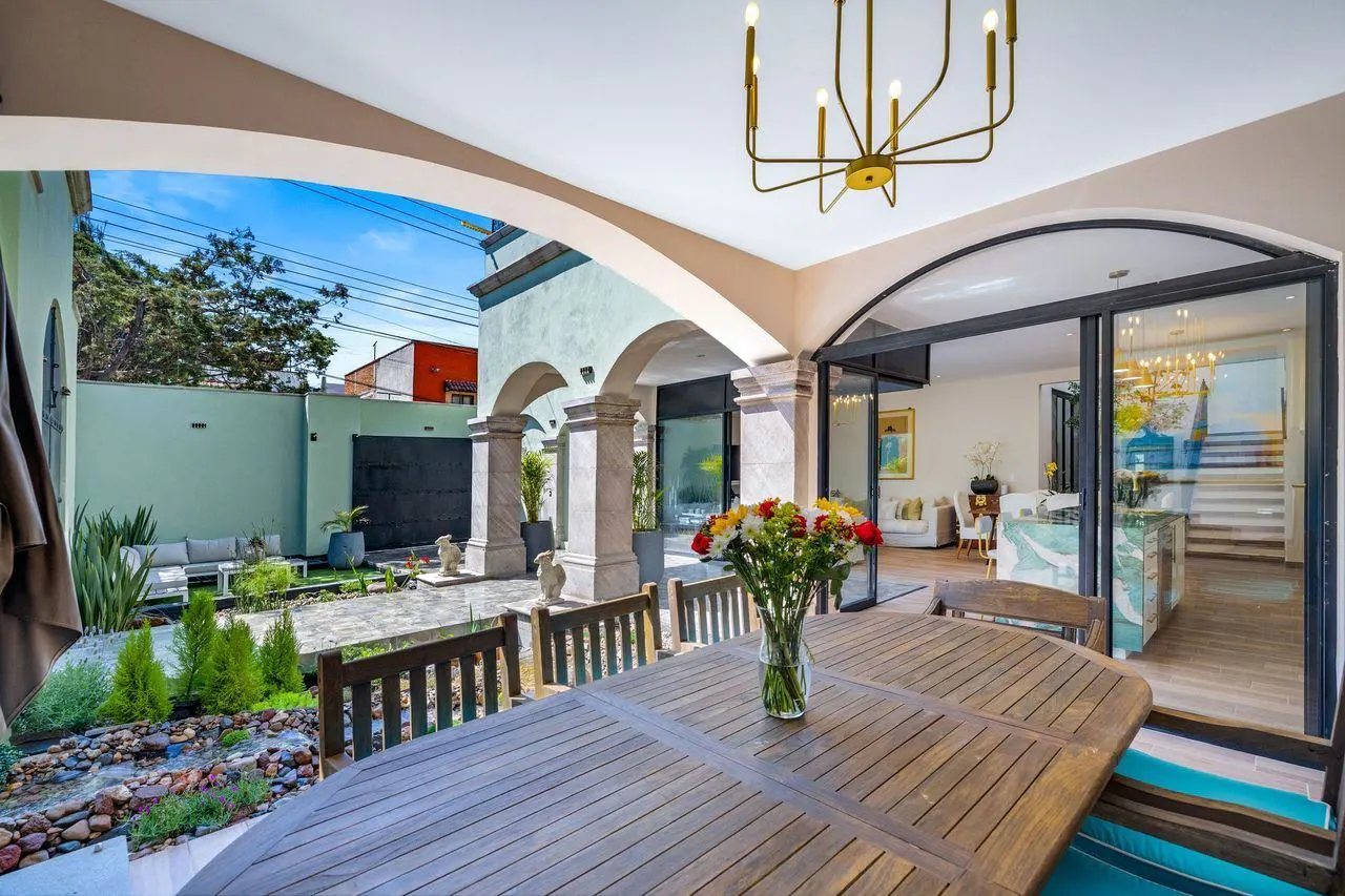 Outdoor patio with a wooden table, chairs, and a vase of flowers. Arched doorway leads to a garden with a green wall and seating. Modern chandelier hangs above.
