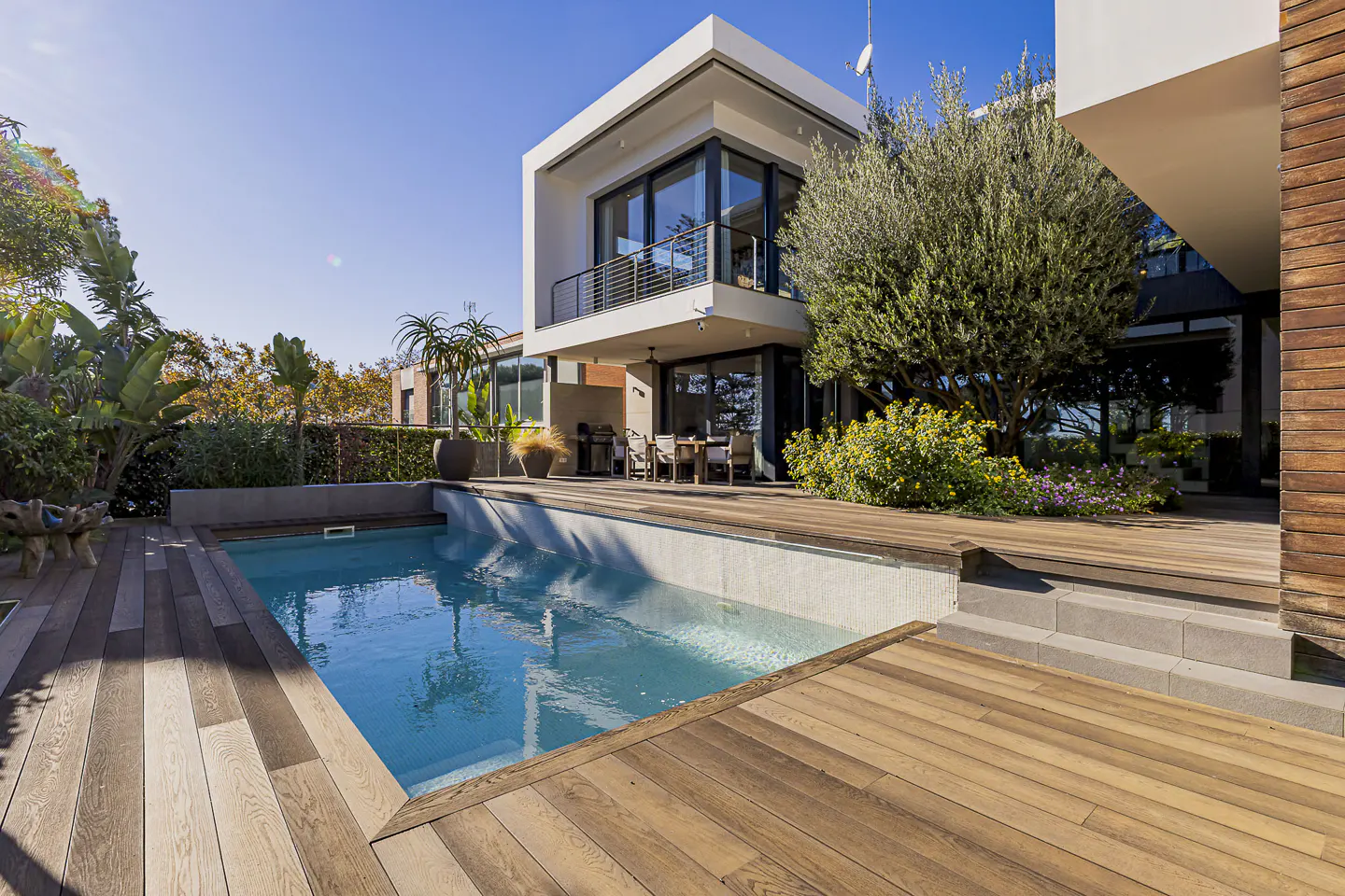Modern home exterior with a blue pool, wood deck, and patio furniture under a balcony on a sunny day.