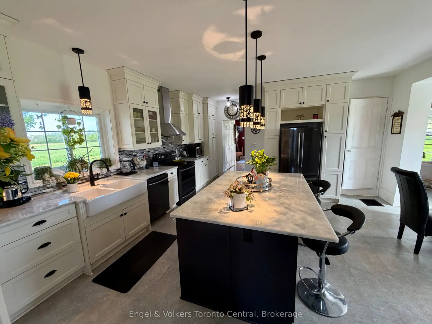 A bright kitchen with white cabinets, a black island with a marble countertop, and pendant lights. A large window overlooks a green field.