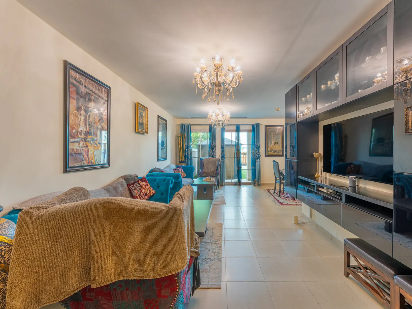 Spacious living room with a brown sofa, teal armchairs, a large wall-mounted TV unit, and two ornate crystal chandeliers.