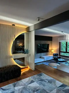 Modern living room with wood floors, a gray patterned rug, and a black ottoman. A lighted, half-circle mirror hangs on a slatted wall. A TV is visible in the background.