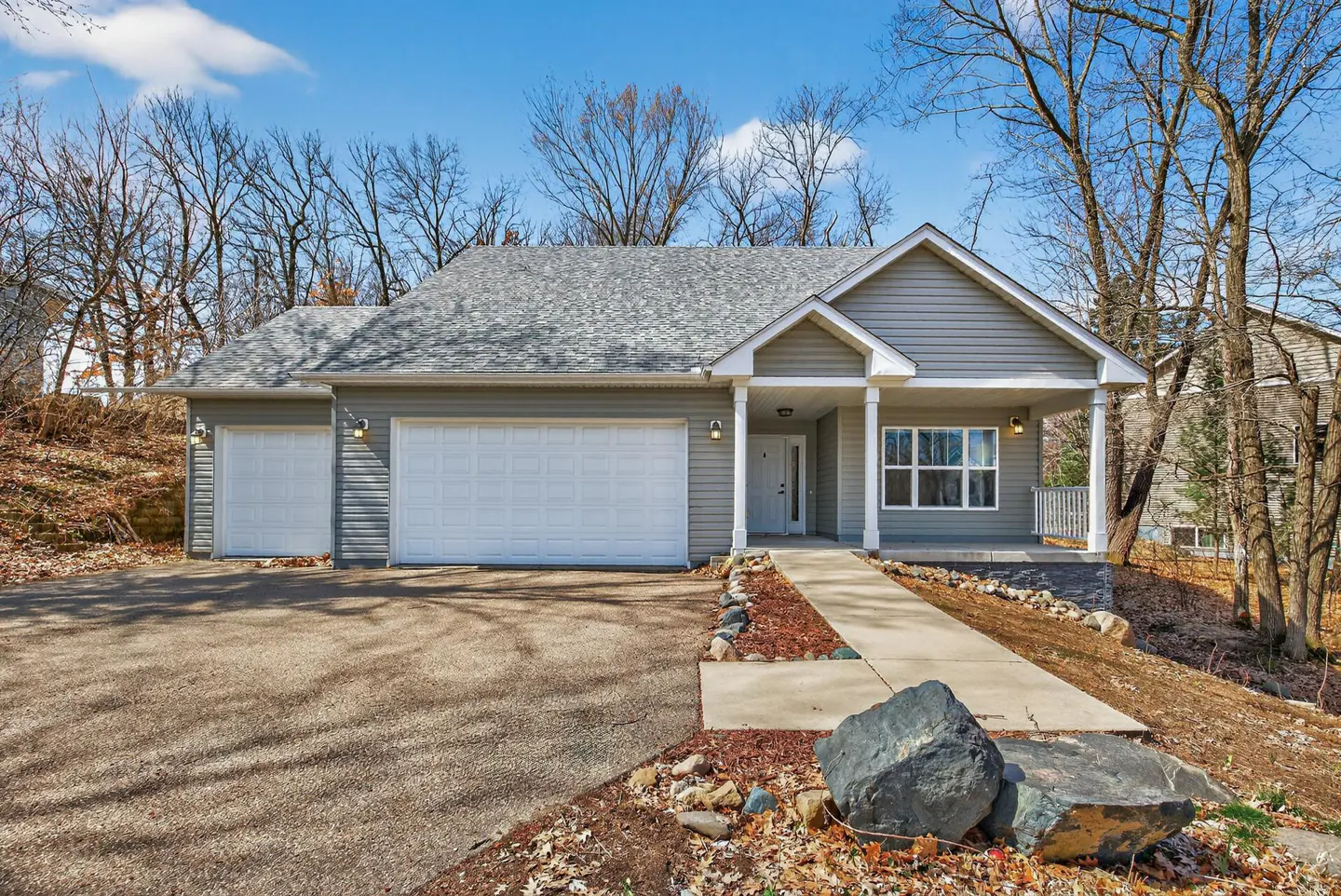 A gray, one-story house with a two-car garage, a gray roof, and a concrete driveway. The house has a small porch with white columns. Trees surround the house.