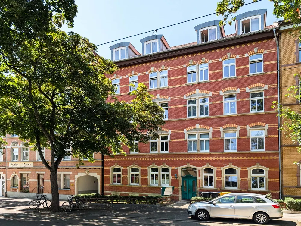 Exterior of a four-story red brick apartment building with white windows and a teal door. A silver car is parked in front.
