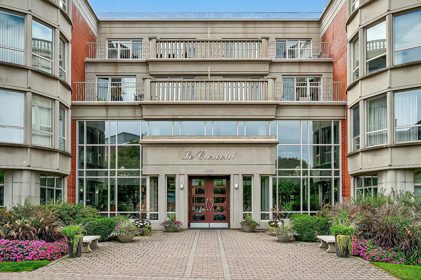 Exterior of "Le Crescent" building with balconies, large windows, and a brick walkway lined with flowers and benches.