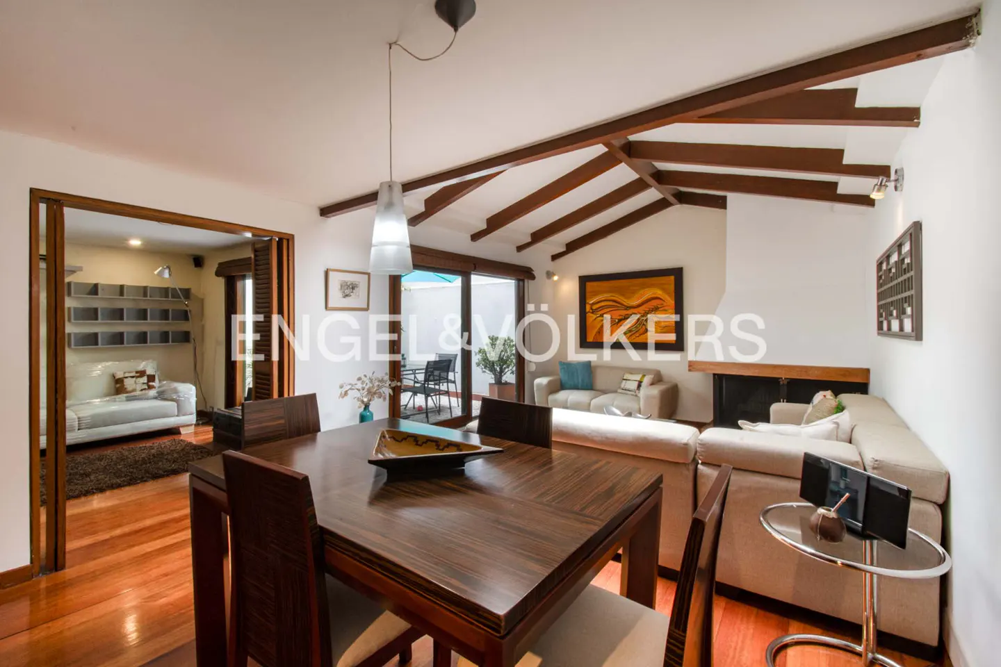A bright, open-concept living and dining area with hardwood floors and exposed beams. A dark wood table sits in the foreground.