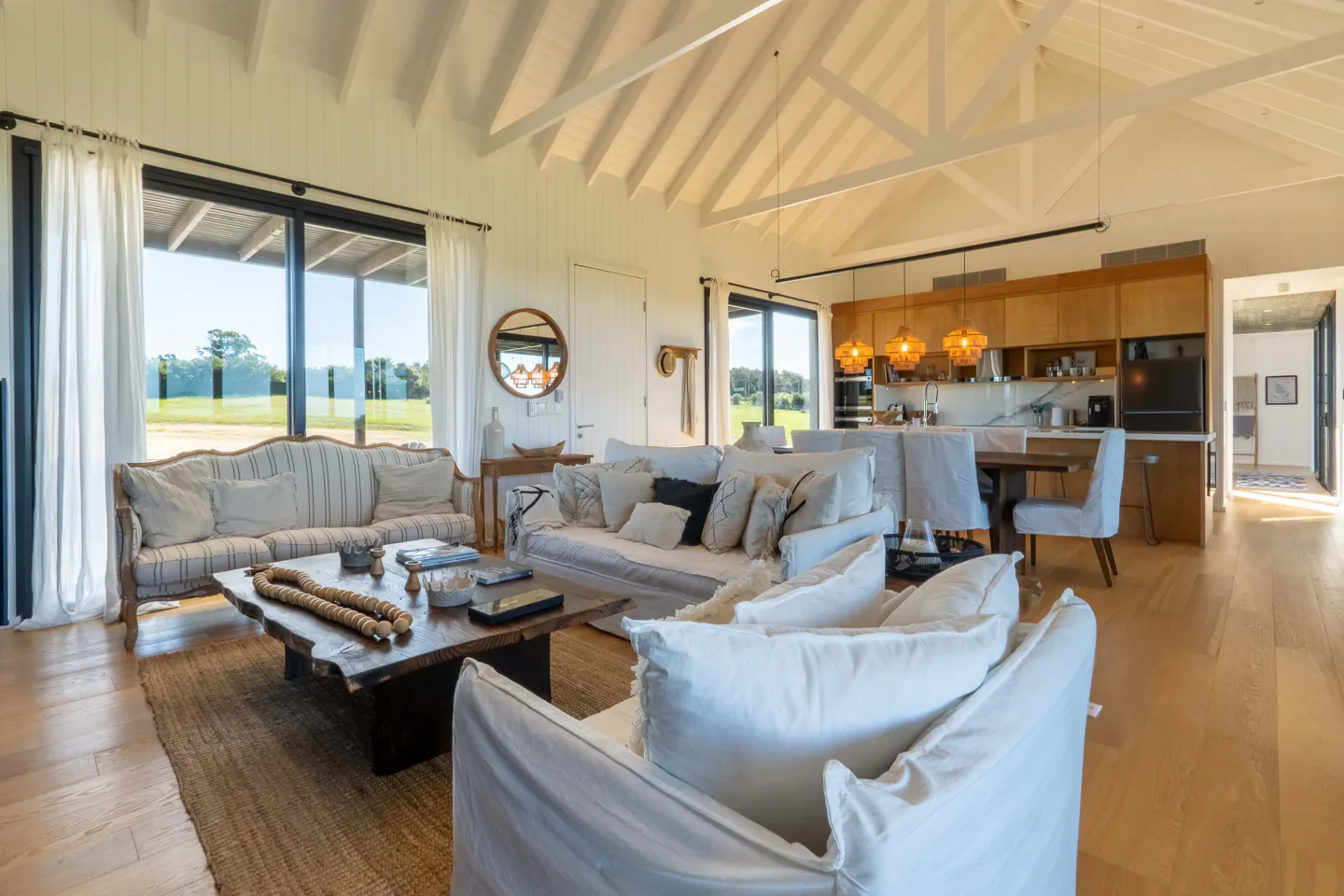 Bright, open-plan living room with white sofas, wood floors, and a rustic coffee table. Large windows offer views of a green landscape.