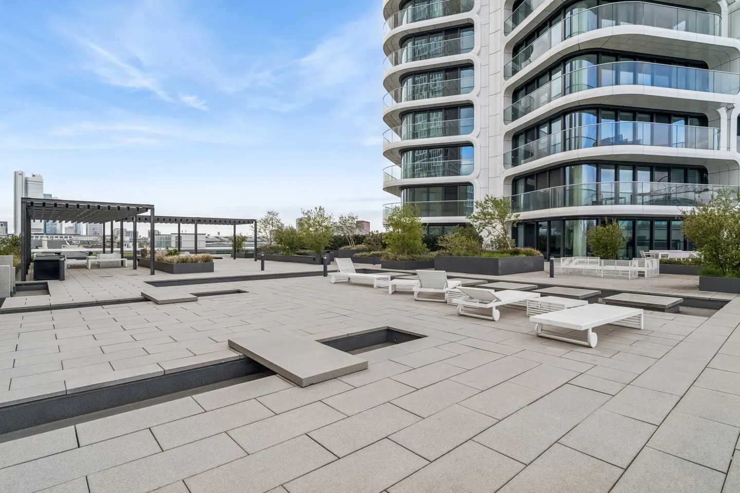 Rooftop patio with white lounge chairs, tables, and a pergola. A modern white building is in the background.