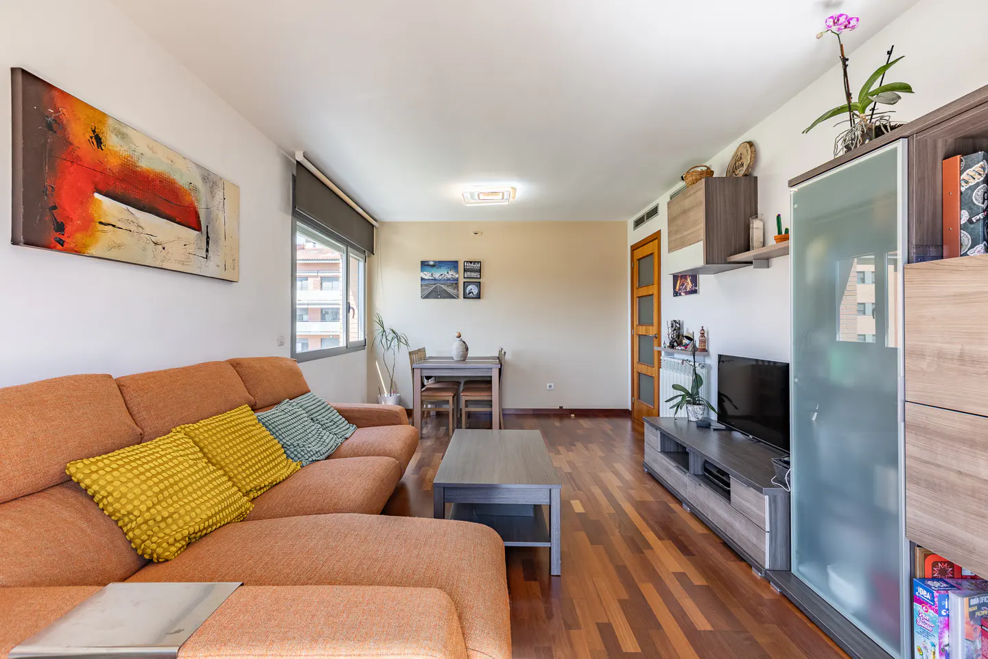 Living room with orange sofa, yellow and blue pillows, wood floors, TV, and abstract art.