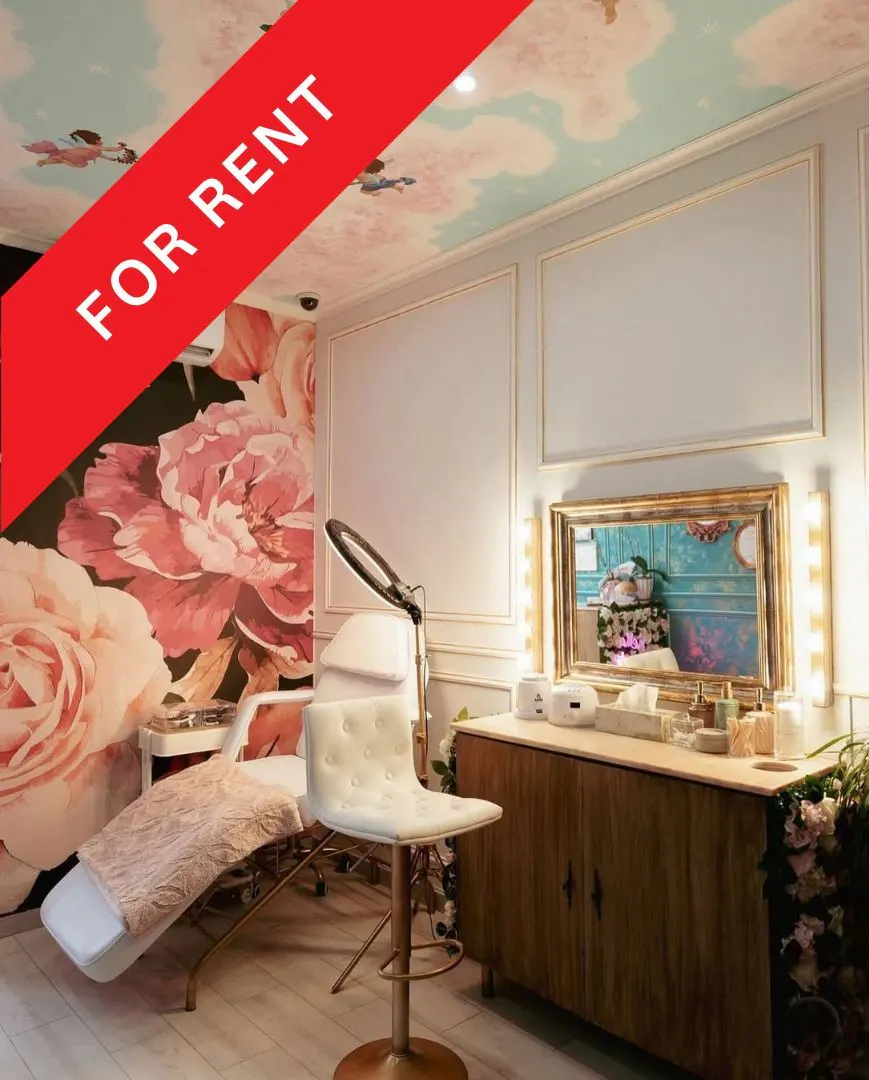 A beauty salon room for rent, featuring a white treatment chair, floral wallpaper, and a vanity with a gold-framed mirror.