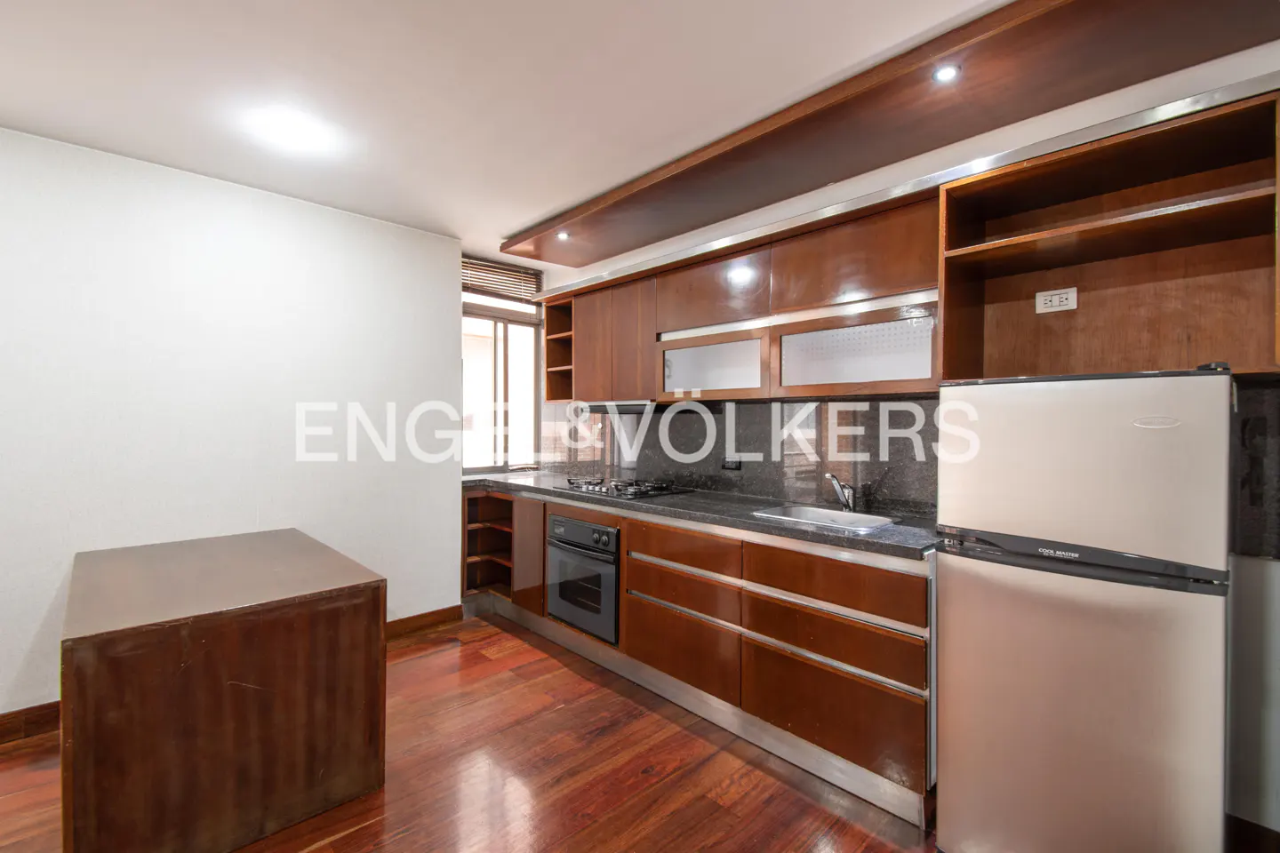 A kitchen with dark wood cabinets, stainless steel refrigerator, and dark countertops. A wood table sits on the wood floor.