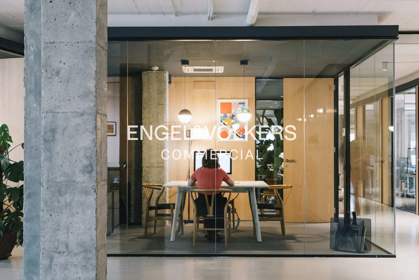 Engel & Volkers office with a person working at a white table. Glass walls, wooden accents, and a concrete pillar add modern style.