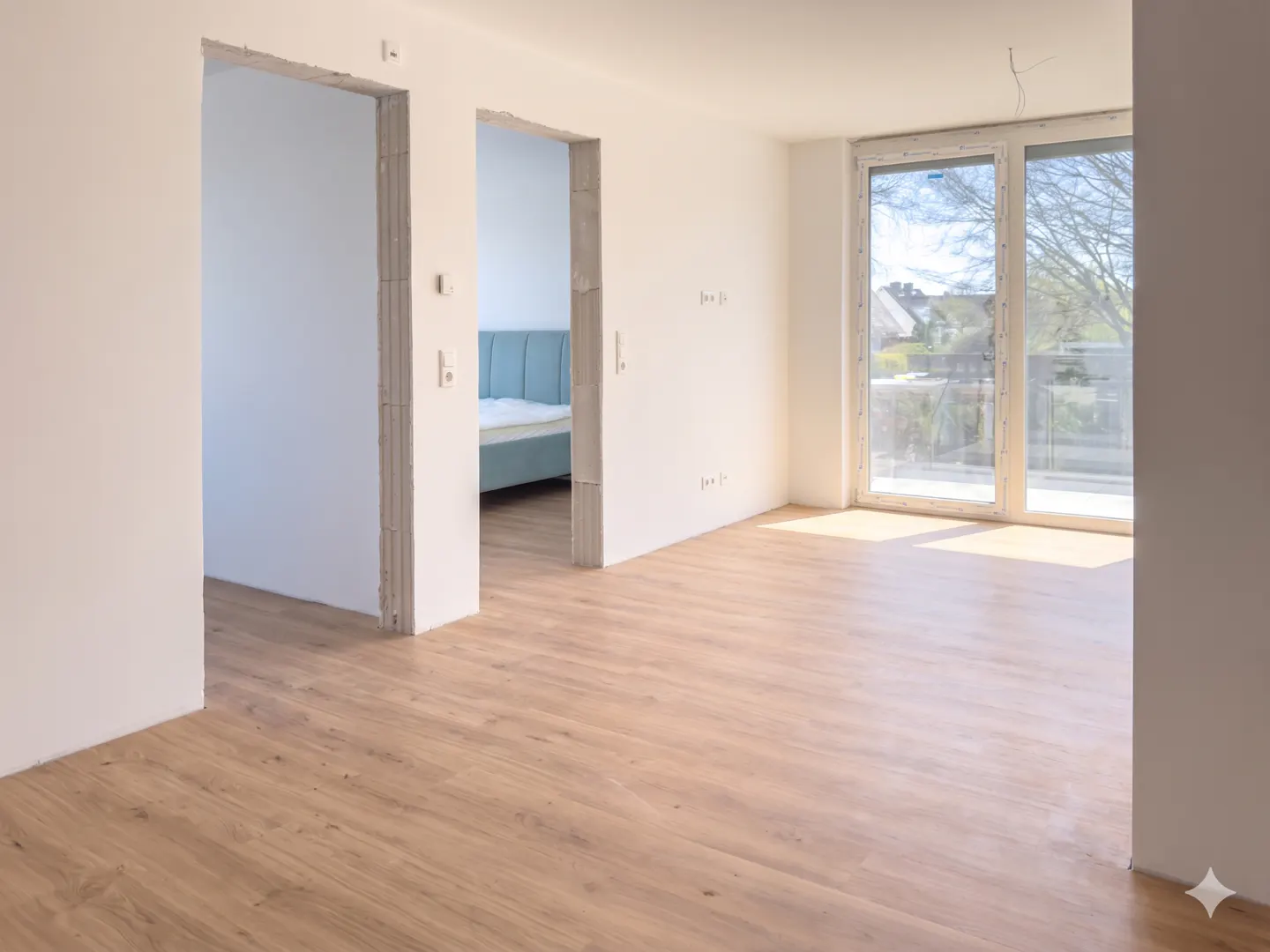 Bright, empty room with light wood floors, white walls, and a large window. A doorway leads to a bedroom with a blue headboard.