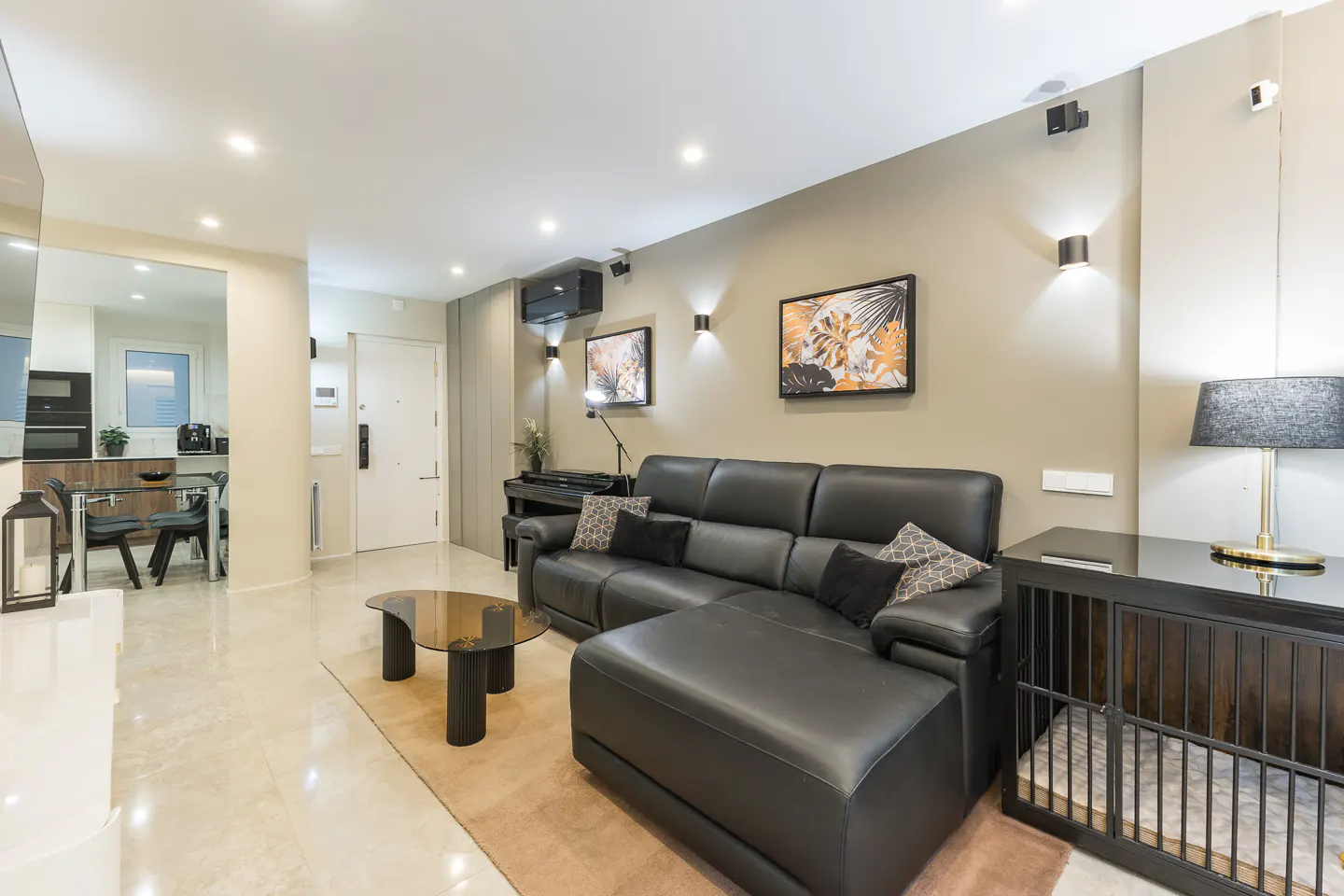 Modern living room with black leather sofa, glass coffee table, piano, and dog crate. Beige walls and marble floors.