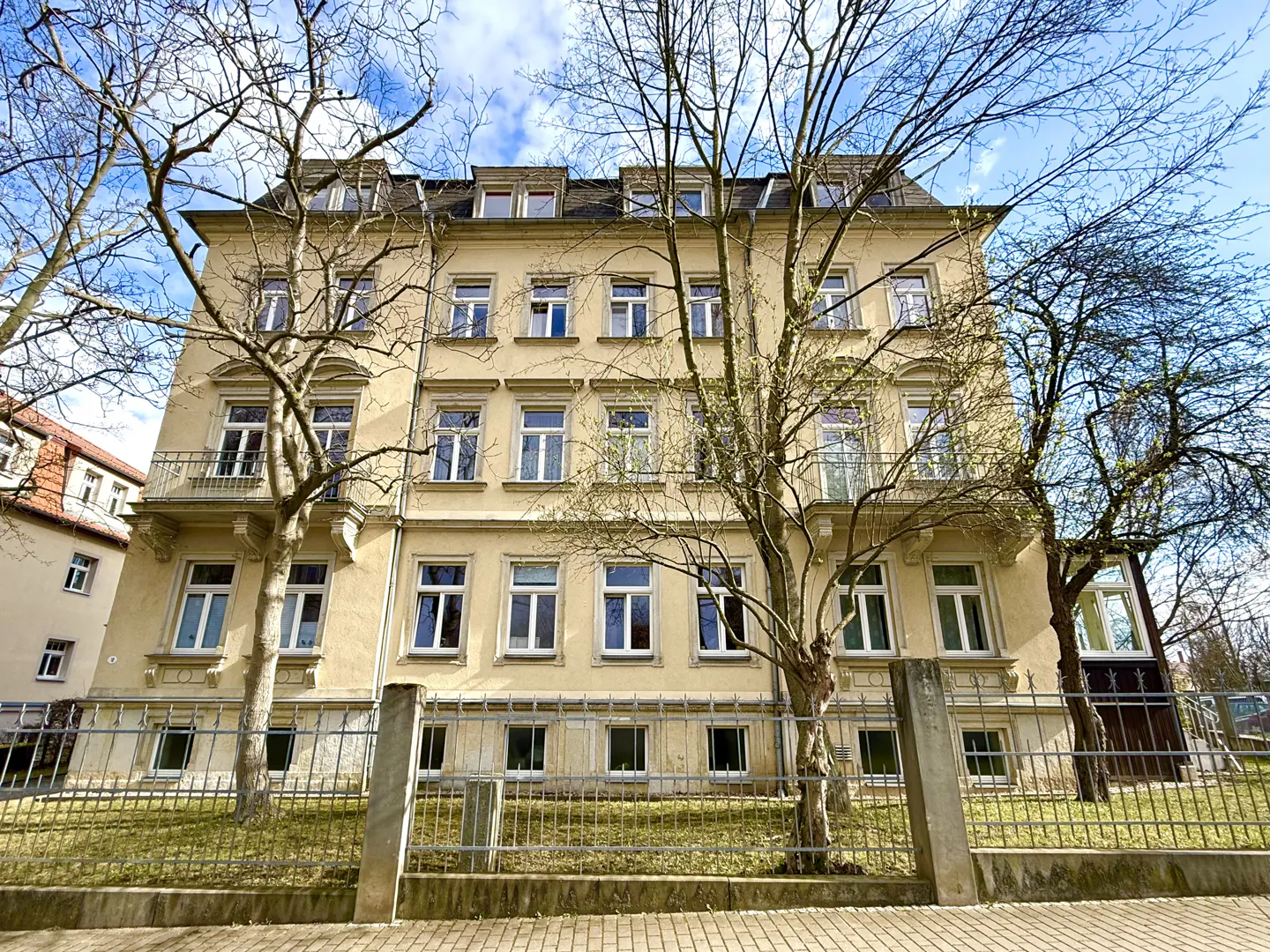 A three-story, light yellow apartment building with many windows and small balconies, framed by bare trees and a metal fence.