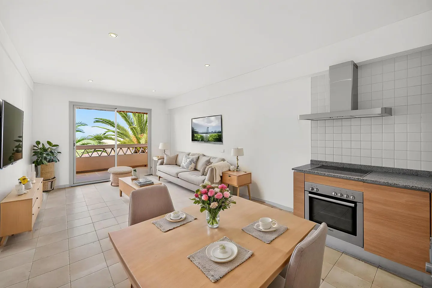 Bright, open-plan apartment with a dining table set for two, a beige sofa, and a balcony with palm trees. The kitchen has wood cabinets.