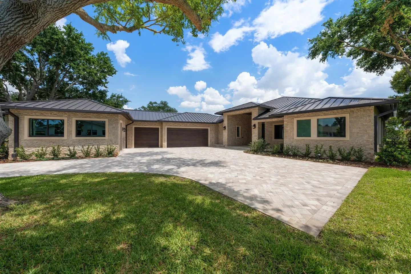 Beige stone house with a dark metal roof, three-car garage, and a paver driveway on a sunny day.