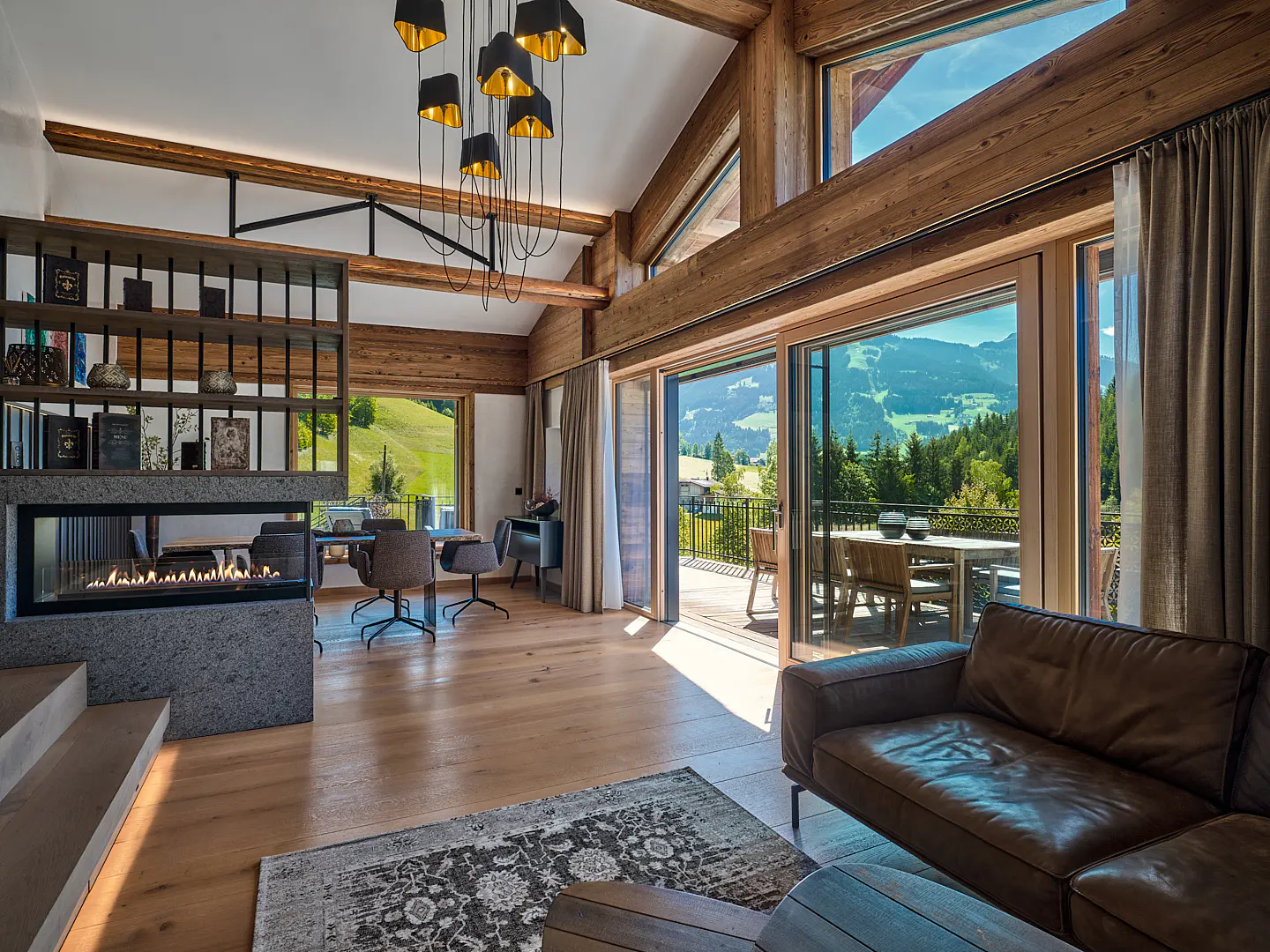 Living room with wood floors, beams, and a stone fireplace. Large windows offer mountain views. A brown leather sofa sits on a patterned rug.
