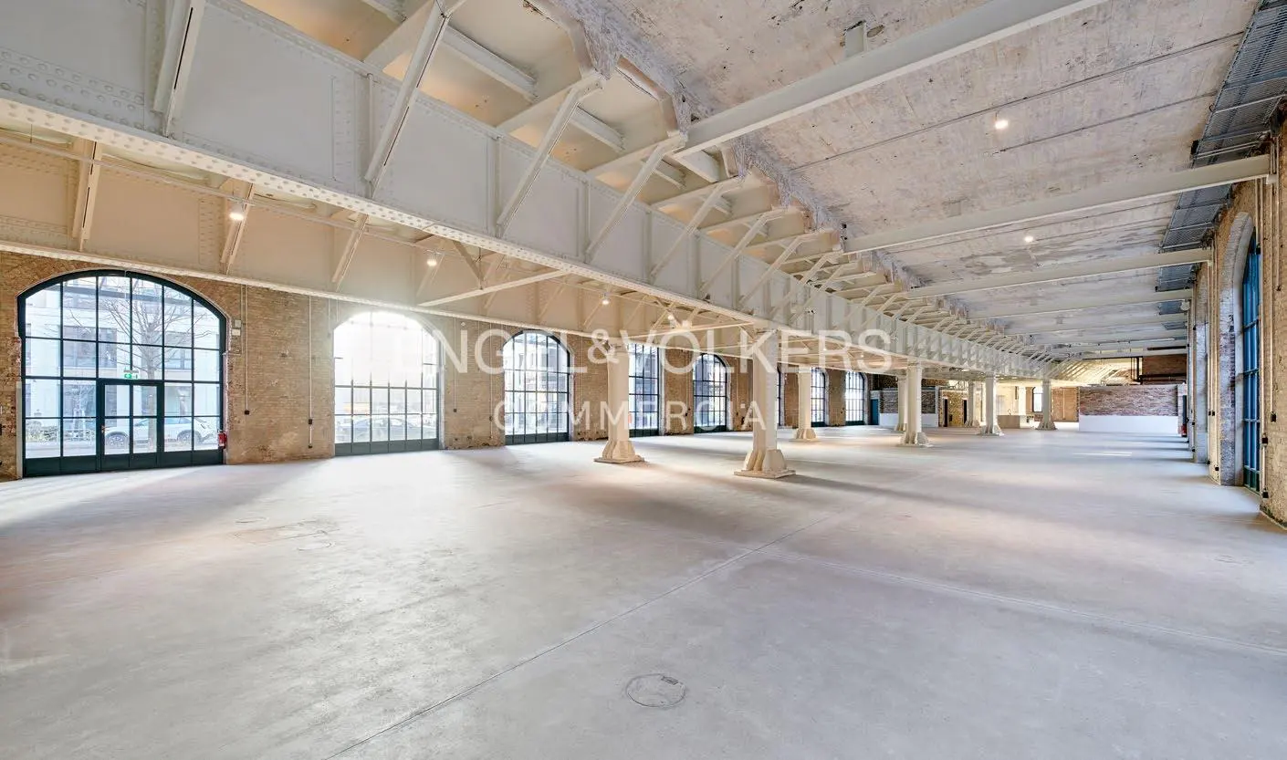 Bright, empty commercial space with concrete floors, brick walls, and large arched windows. White steel beams support the ceiling.