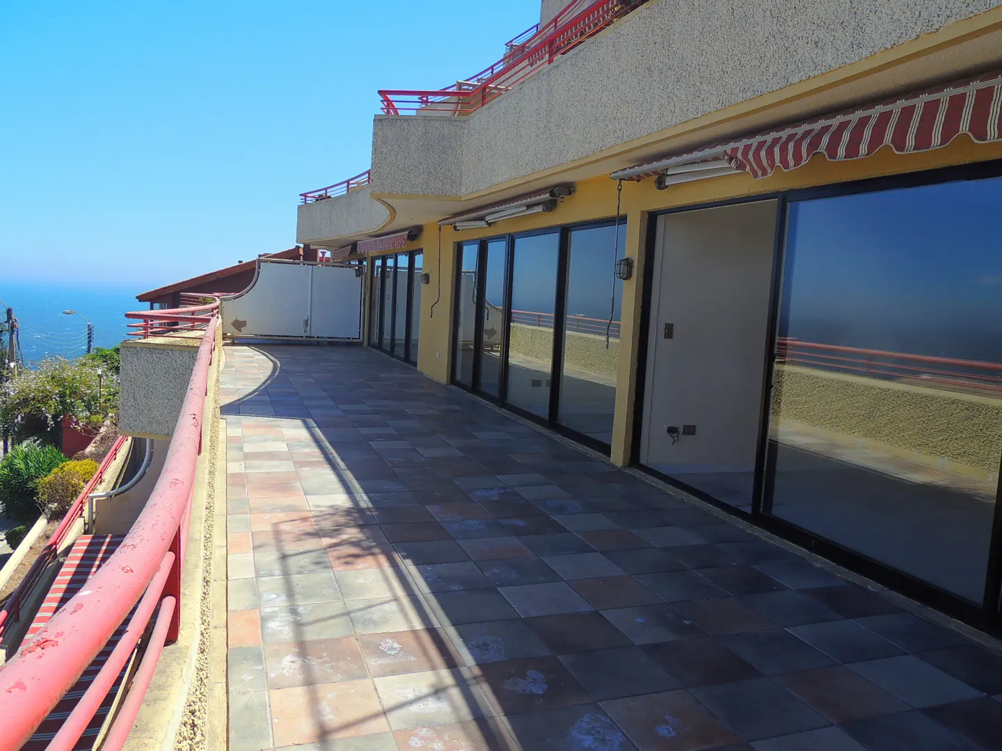 A long, tiled balcony with a red railing overlooks the ocean. Large glass doors reflect the view.