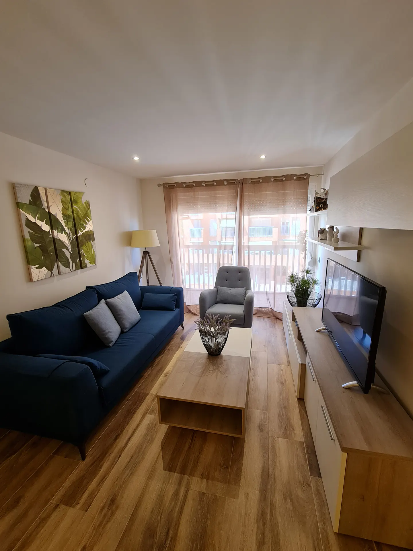 Living room with blue sofa, gray armchair, wood floors, and a TV. A tropical leaf painting hangs above the sofa. Curtains cover the balcony doors.