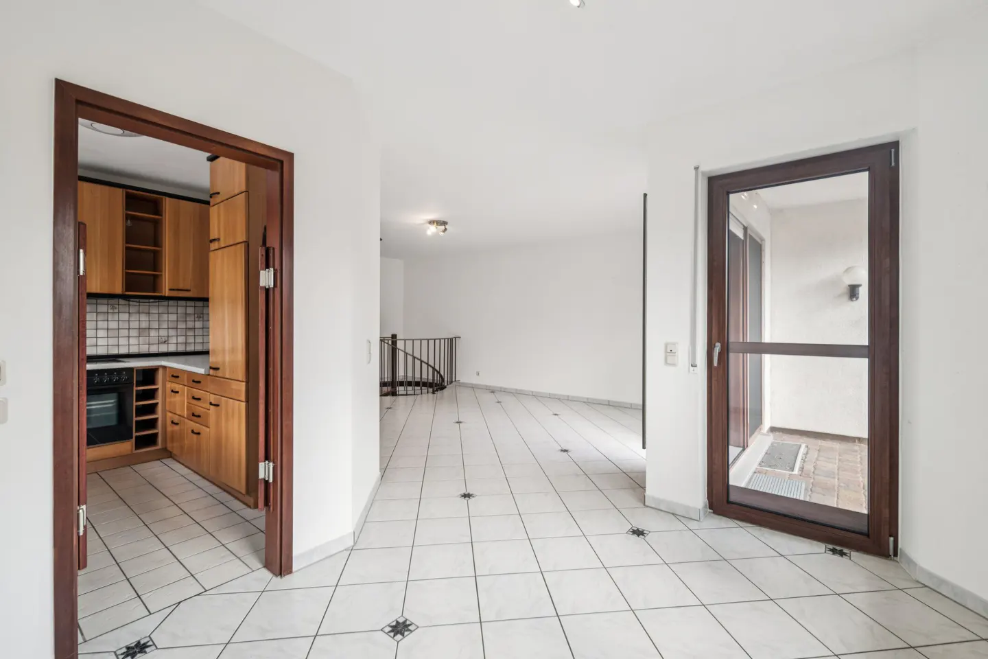 Bright hallway with white tiled floors, a wooden-framed kitchen doorway, and a glass door leading to a balcony.